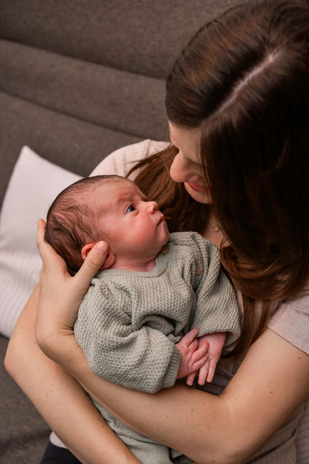 Eine  sitzende Mutter mit langen braunen Haaren hält ihr Baby im grünen Strampler auf dem Arm und beide schauen sich in die Augen.